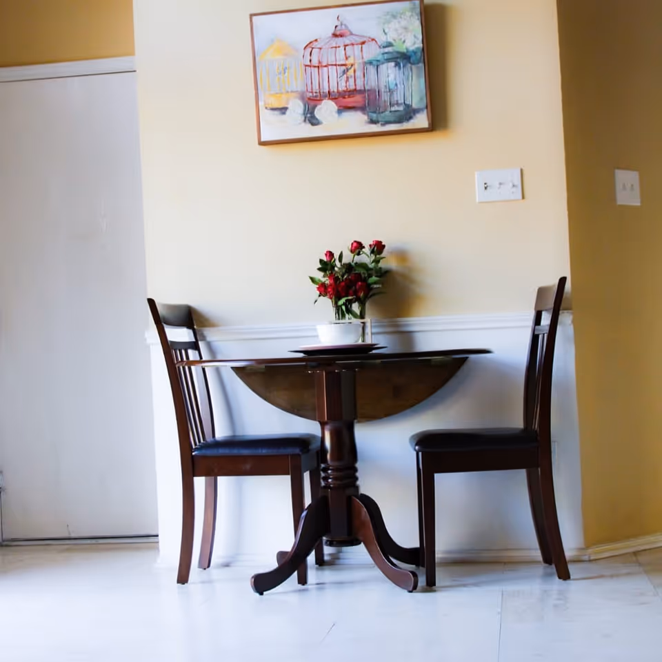A small wooden dining table with a vase of red flowers in the center, flanked by two wooden chairs with dark cushions, set against a beige wall with a painting of colorful birdcages hanging above.