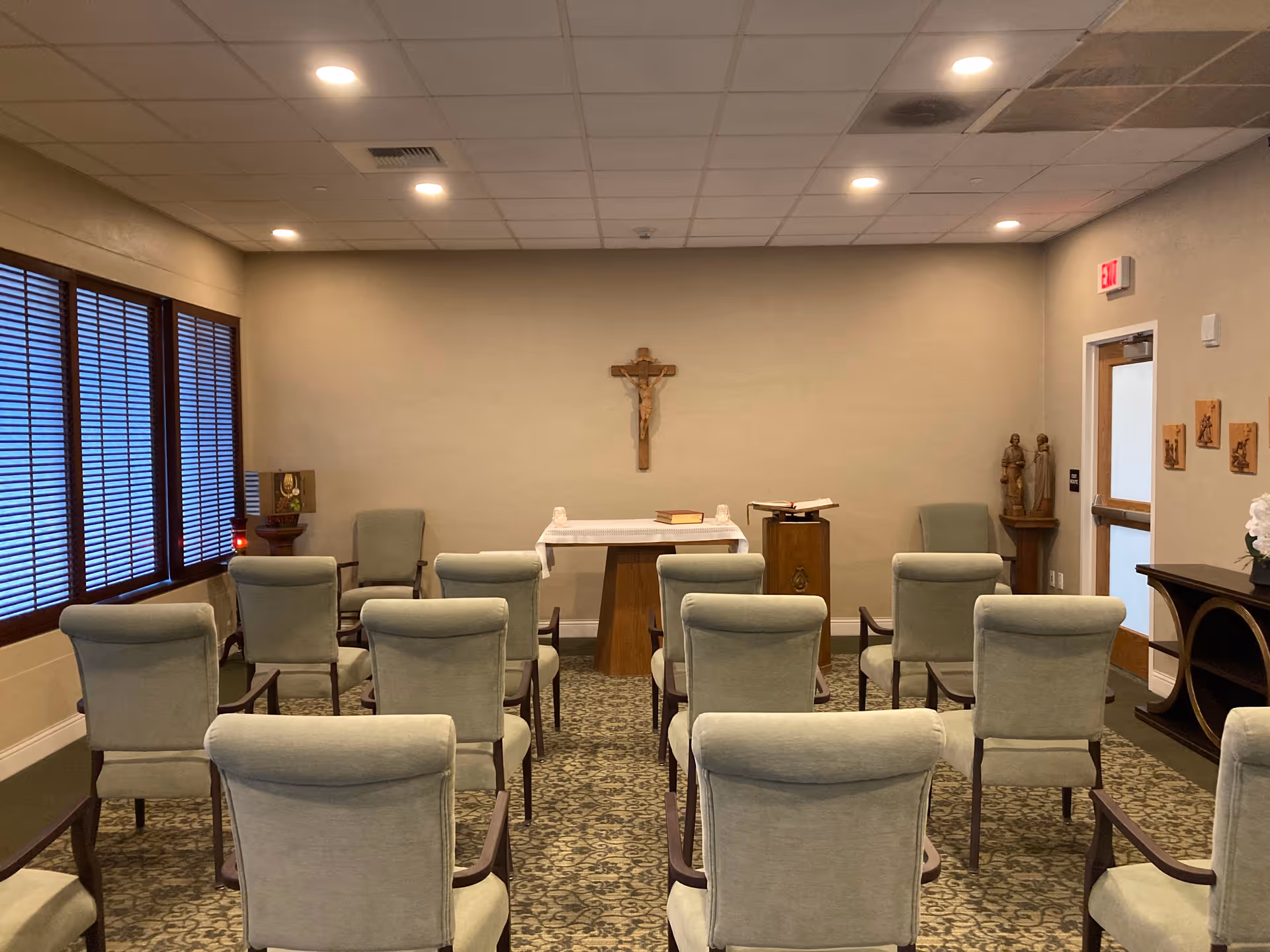 A small chapel room with rows of light green upholstered chairs facing a wooden altar table covered with a white cloth. A crucifix is mounted on the beige wall behind the altar. The room has a patterned carpet, wooden window blinds on the left, and religious statues and plaques on the right wall near an exit door.
