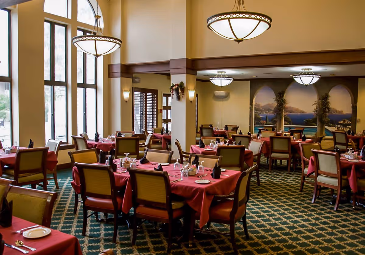 A spacious dining room with multiple tables covered in red tablecloths, each set with plates, glasses, and black napkins. The room features large windows allowing natural light, elegant hanging light fixtures, and a mural on the far wall depicting an outdoor scenic view with arches and greenery.