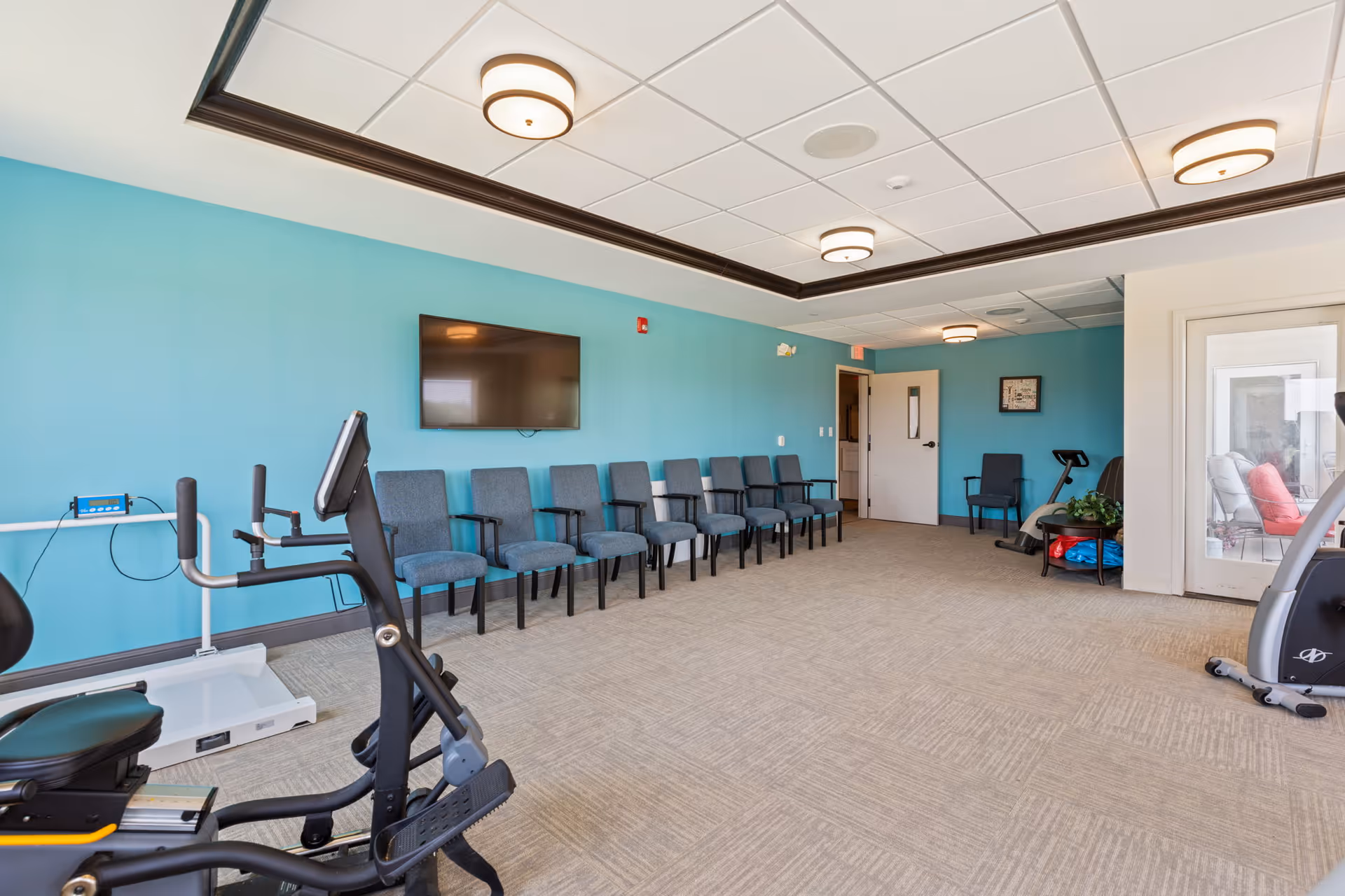 Bright interior exercise/multipurpose room with cardio equipment, a row of chairs, and a wall-mounted TV.