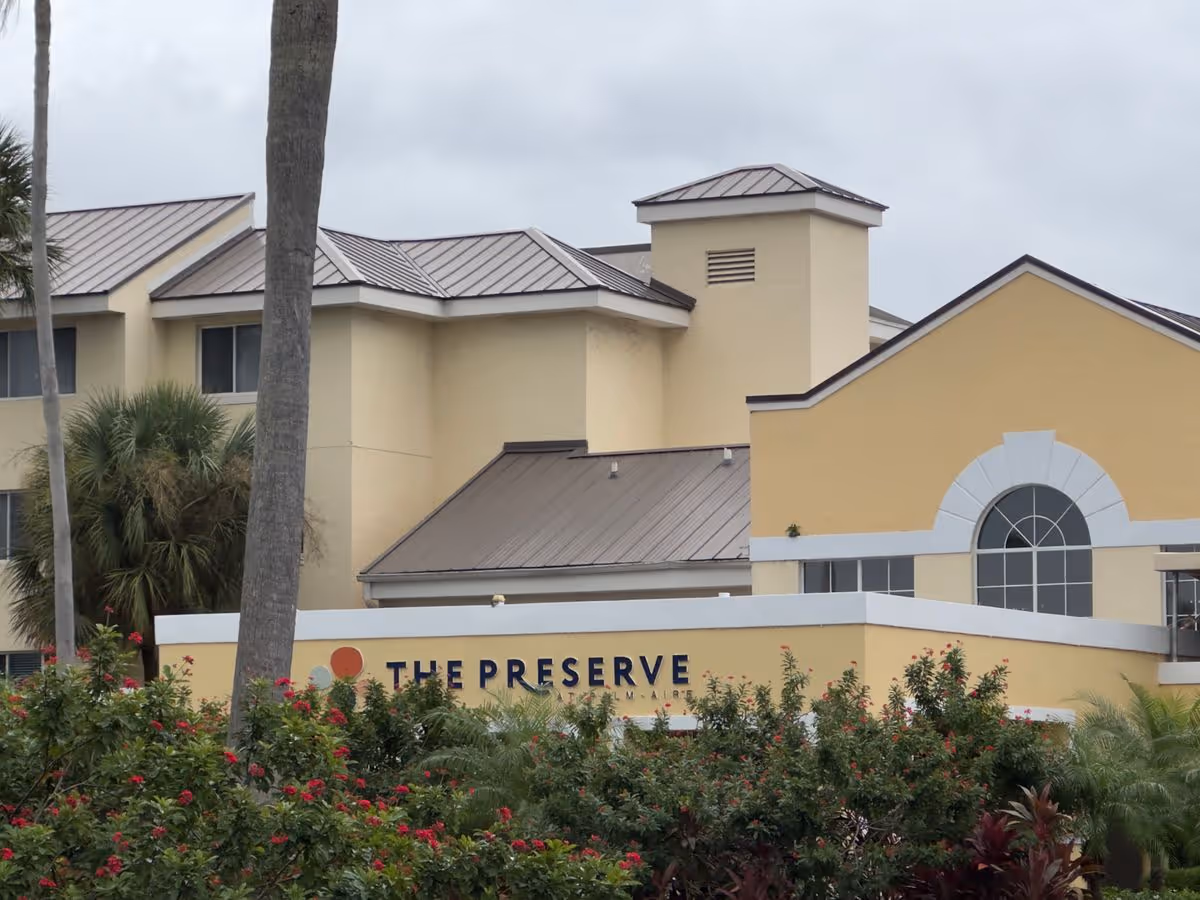 Front exterior of The Preserve at Palm Aire building with a yellow facade, arched window, metal roofs, and landscaped shrubs.