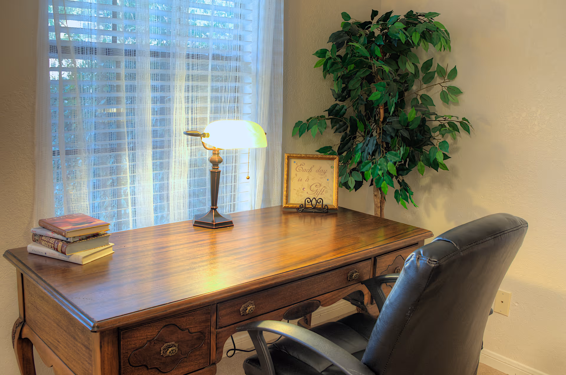 A wooden desk by a window with a lamp, a stack of books, a framed sign, a potted plant, and a black leather chair.