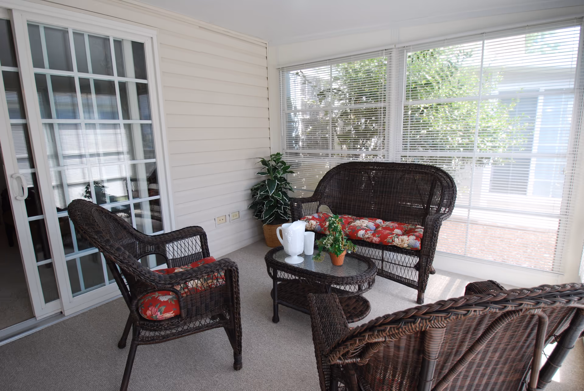 A sunroom with wicker furniture including a loveseat and two chairs with red floral cushions, a glass-top coffee table with a white pitcher and cups, and a small potted plant. The room has large windows with blinds and a sliding glass door leading to an interior space.