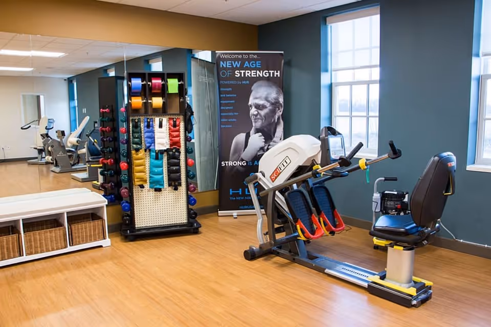 A bright indoor fitness room with a seated exercise machine, weight rack, colorful resistance bands and large windows.