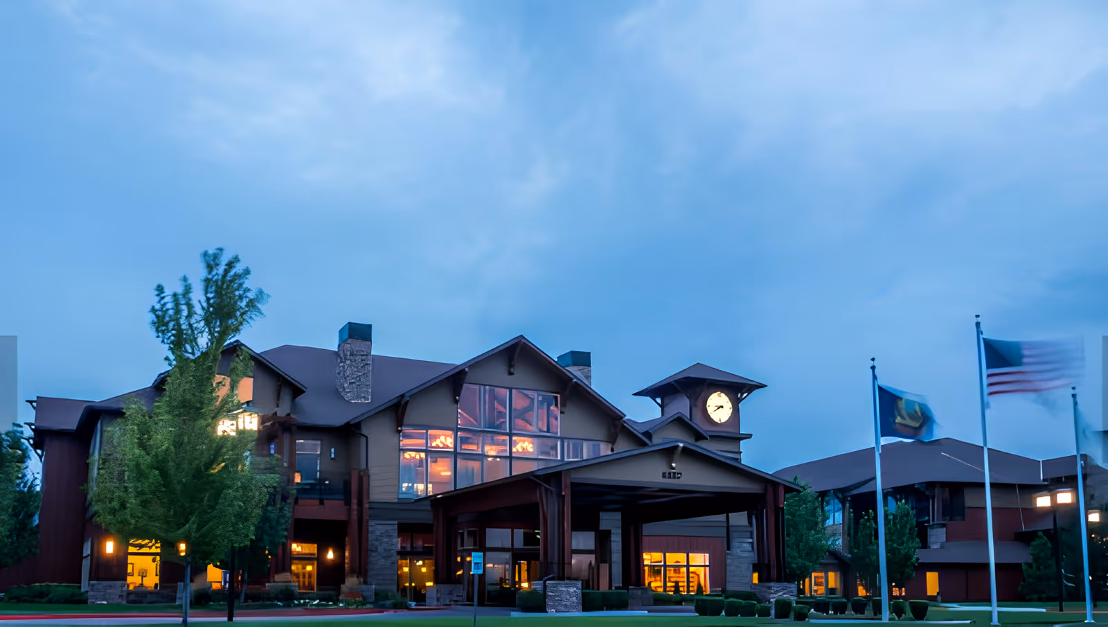 Front exterior of a large senior living facility at dusk with illuminated windows, a clock tower, and flags in front.
