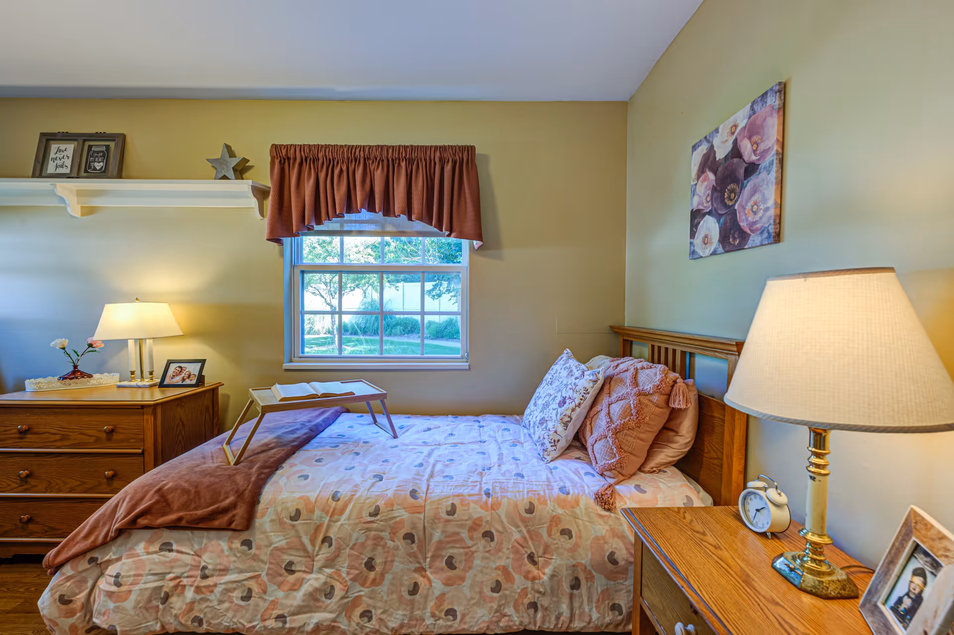 A cozy bedroom with a single bed covered in a floral patterned comforter and multiple pillows. There is a wooden nightstand on each side of the bed, each with a lamp and framed photos. A window with a red valance lets in natural light, and a floral painting hangs on the wall above the bed. A wooden dresser with a lamp, flowers, and framed photos is positioned against the wall under a white shelf holding decorative items.