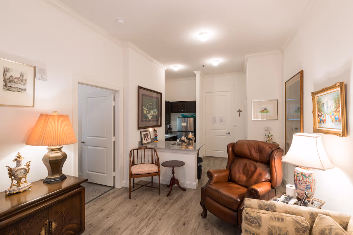 A cozy living area in a senior living facility featuring a brown leather armchair, a patterned sofa, a wooden side table with a decorative lamp, and a small kitchen area with granite countertops and dark cabinets. The walls are adorned with framed artwork, and the flooring is light wood. There is a door leading to another room and a white entrance door in the background.
