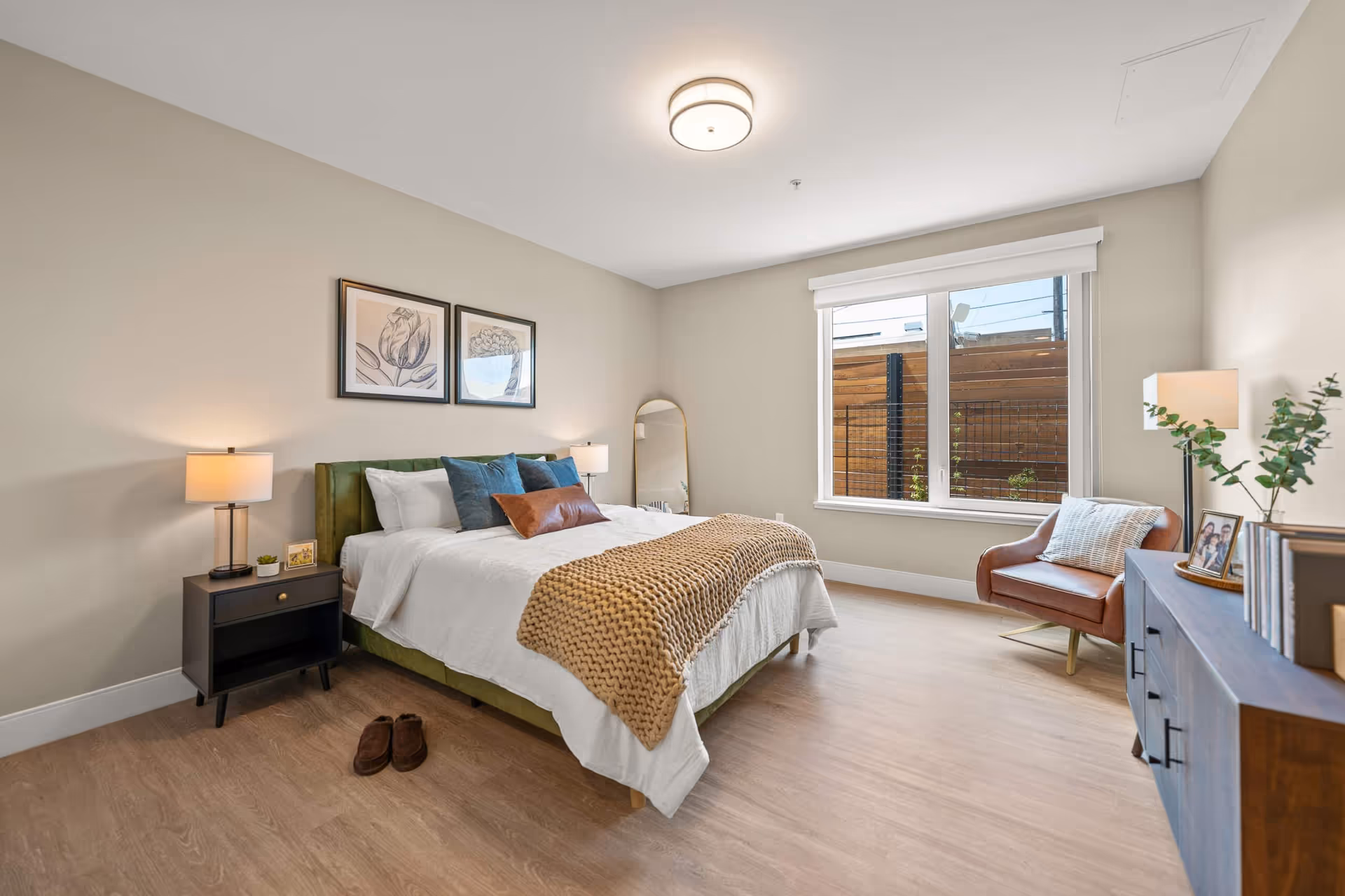 A bright and modern bedroom with a large bed featuring white bedding, blue and brown pillows, and a beige knitted throw. There are two framed floral artworks above the bed, a nightstand with a lamp and small decor items, a full-length mirror leaning against the wall, a window with a view of a wooden fence, a brown leather armchair with a pillow, and a wooden dresser with books, a lamp, and a plant.