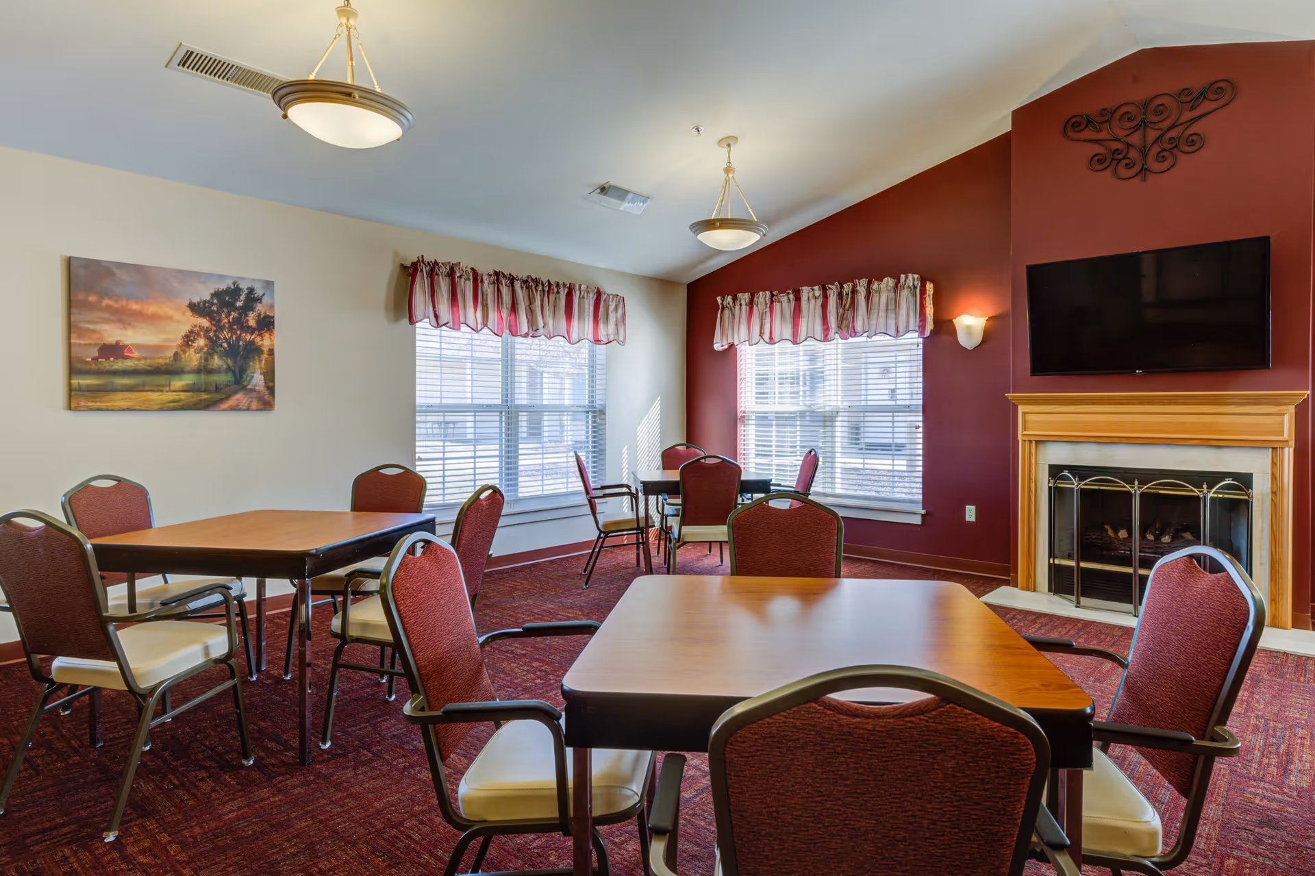 A cozy senior living community common area with several tables and red cushioned chairs arranged on a red carpet. The room features large windows with red and white valance curtains, a fireplace with a wooden mantle, and a flat-screen TV mounted above it. The walls are painted in a combination of beige and dark red, and there is a landscape painting on one wall.