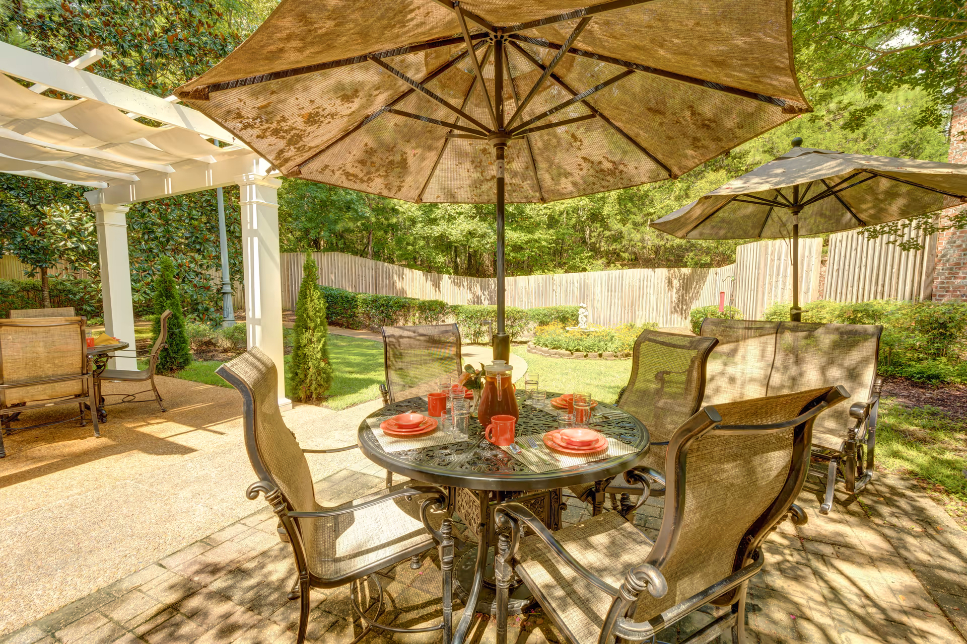 Outdoor patio area with a round metal table set for four with red plates, mugs, and glasses. The table is shaded by a large umbrella. There are additional chairs and another umbrella nearby. The patio is surrounded by greenery, a wooden fence, and a pergola on the left side.