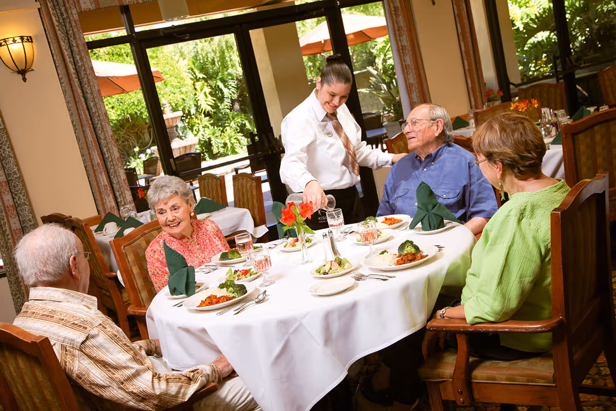 Four elderly people sitting around a round dining table with white tablecloth, green napkins, and plates of food. A waitress is pouring water into a glass while smiling and interacting with the group. The dining room has large windows showing greenery outside.