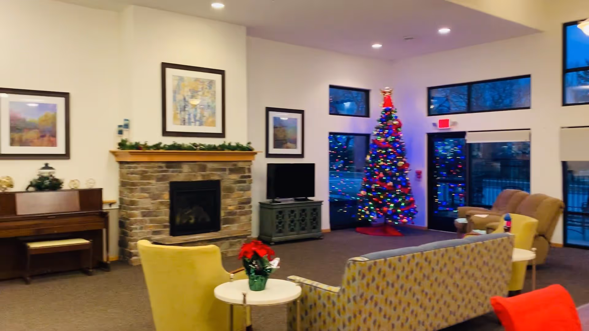 A cozy senior living facility common area decorated for the holidays with a Christmas tree adorned with colorful lights. The room features a stone fireplace with framed artwork above it, a piano on the left, a TV on a stand, and comfortable seating including armchairs and a patterned sofa. Large windows and glass doors provide a view of the outside.