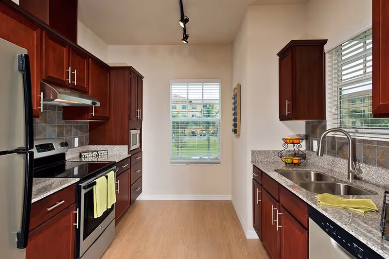 A modern kitchen with dark wood cabinets, stainless steel appliances including a refrigerator, stove, microwave, and dishwasher. The kitchen has granite countertops, a double sink, a window with white blinds, and a light wood floor. There is a fruit basket on the counter and a yellow towel hanging on the oven handle.