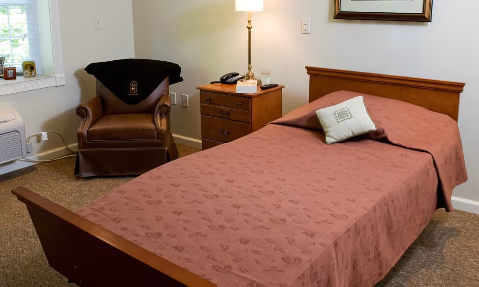A small bedroom with a single bed covered in a patterned red bedspread, a nightstand with a lamp and phone, and a leather armchair by the window.