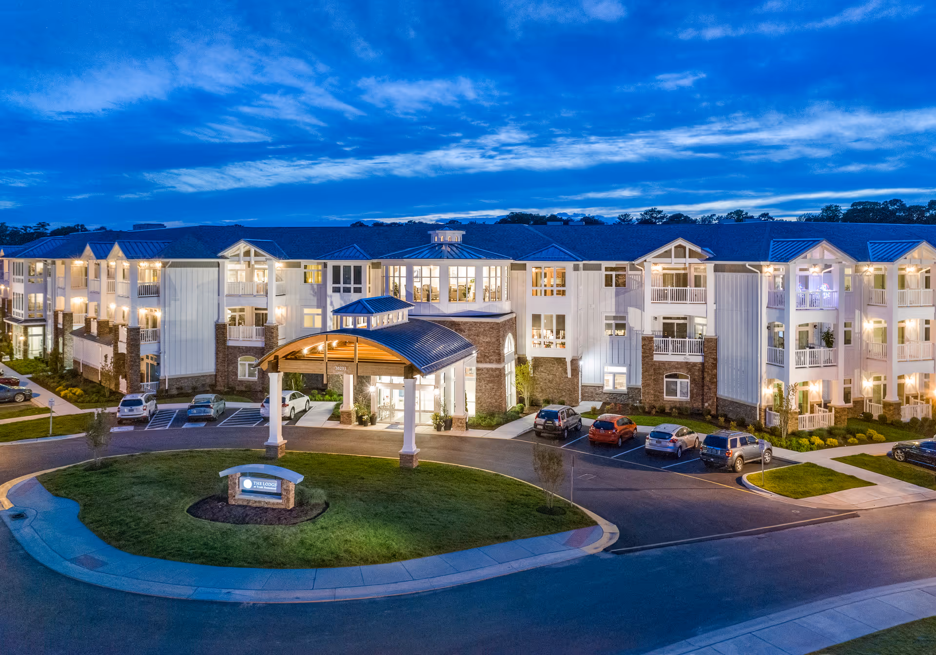 Front exterior of a three-story senior living building at dusk with a covered porte-cochère, landscaped roundabout and parked cars.