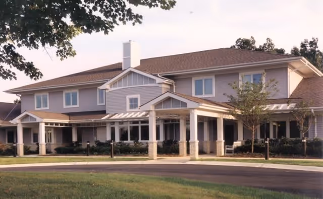 Exterior view of a two-story senior living facility building with a covered entrance, surrounded by trees and landscaping under a clear sky.