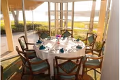 A round dining table set for six people with white tablecloth, teal napkins, and glassware in a sunlit room with large windows overlooking a green lawn and water view.