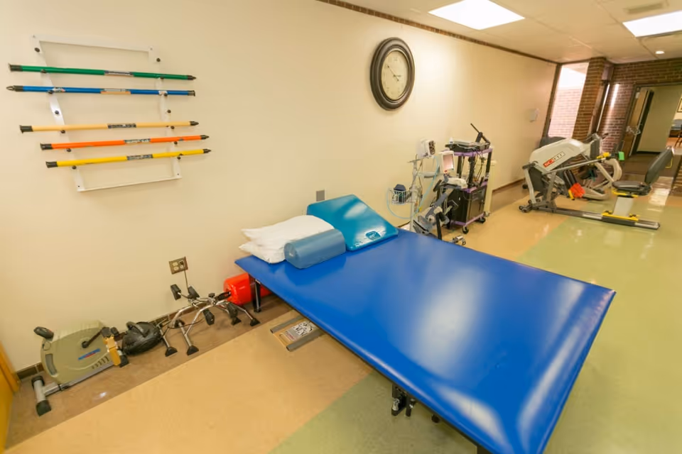 Physical therapy room with a blue treatment table, exercise equipment, and colorful therapy rods mounted on the wall.