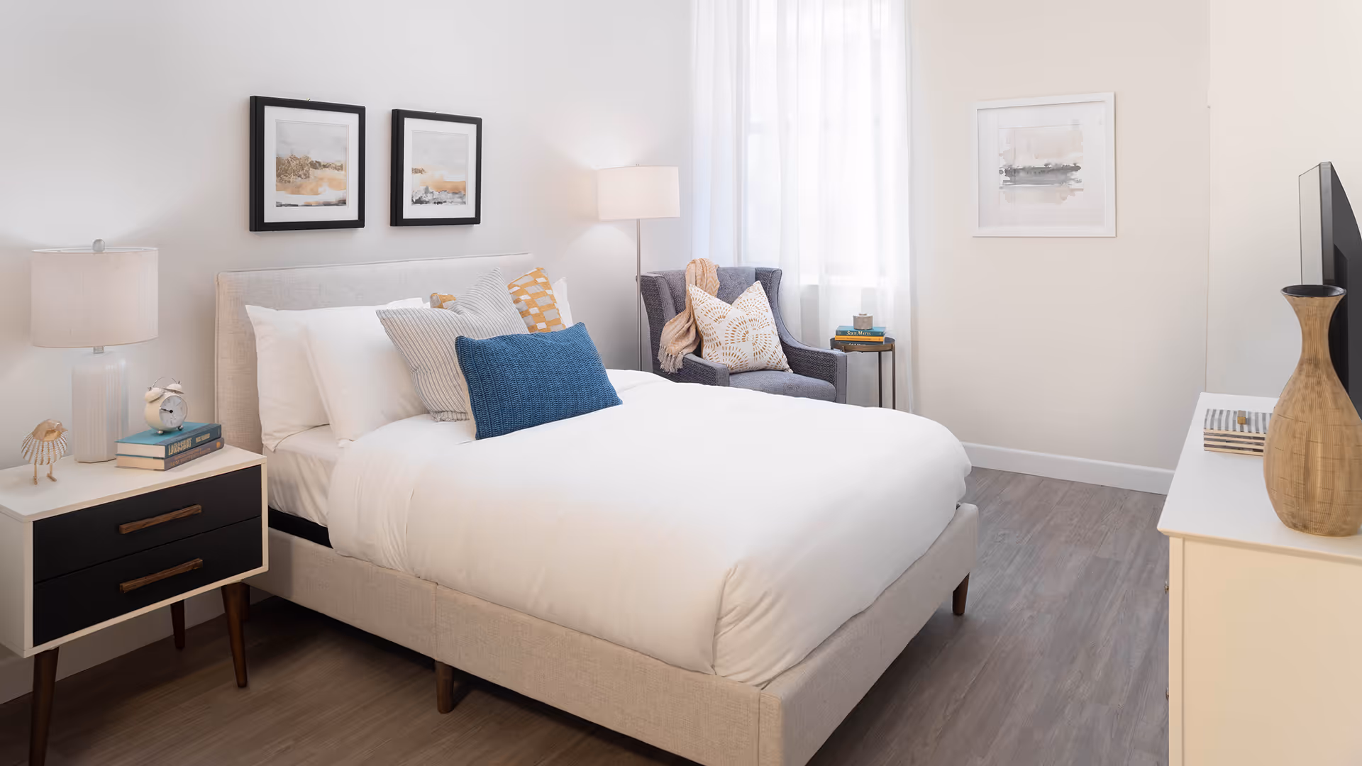A bright and cozy bedroom featuring a neatly made bed with white linens and multiple decorative pillows in blue, gray, and yellow patterns. To the left of the bed is a nightstand with a lamp, an alarm clock, books, and a small decorative bird. On the right side near a window with sheer white curtains, there is a gray armchair with a patterned pillow and a throw blanket, next to a small round side table holding books and a cup. A dresser with a wooden vase and a TV is visible on the right side of the room. The walls are light-colored with framed abstract artwork.