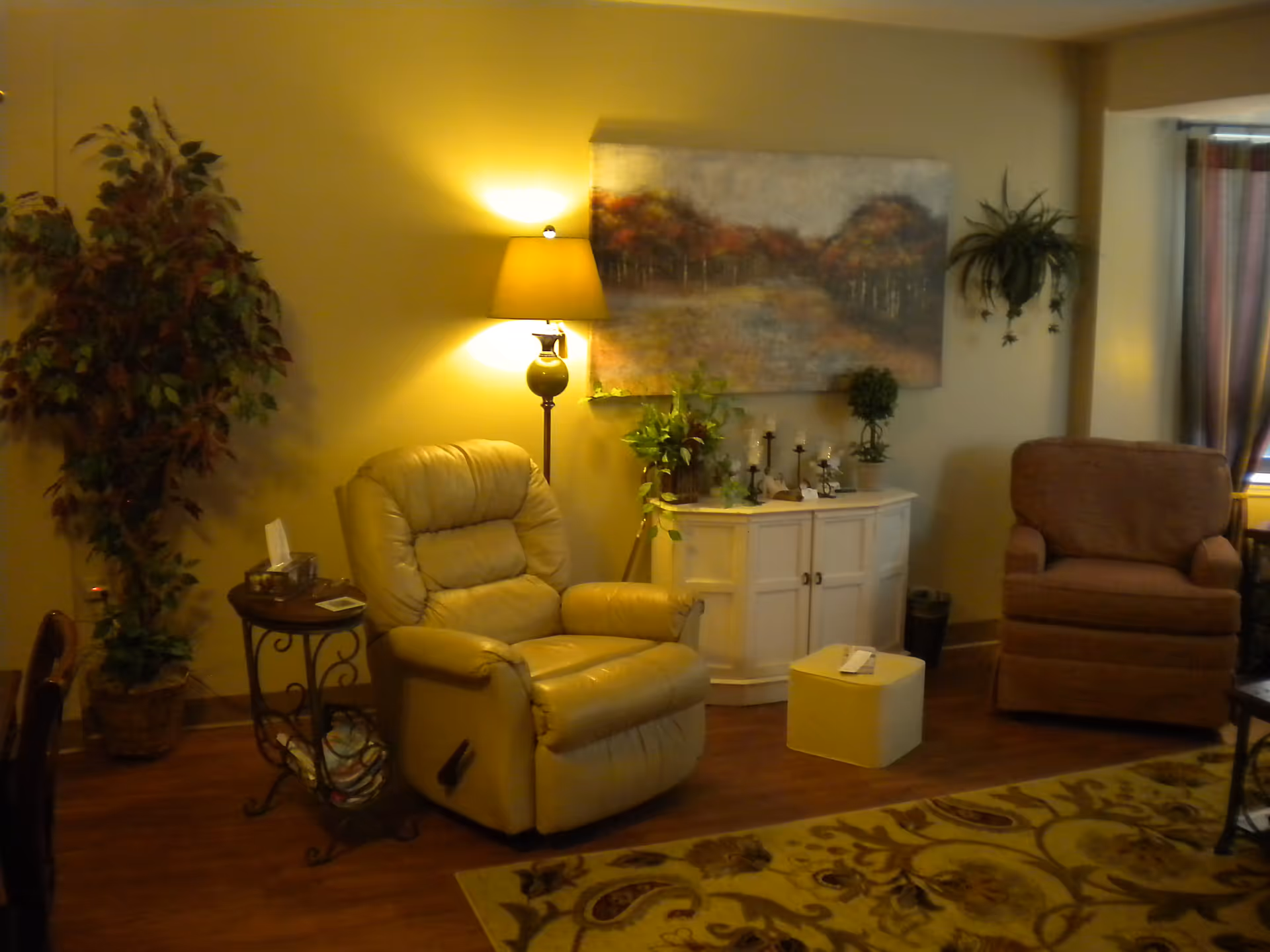 A cozy living room area with a beige recliner chair, a brown armchair, a small white ottoman, and a white cabinet decorated with plants and candles. A floor lamp with a yellow shade illuminates the space, and a large painting of a forest scene hangs on the wall. There is a patterned rug on the wooden floor and a large potted plant in the corner.