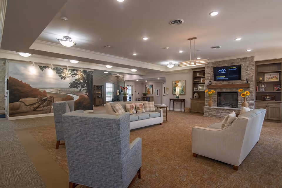 A spacious and well-lit living room area in a senior living facility featuring comfortable sofas and armchairs arranged around a stone fireplace with a mounted flat-screen TV above it. The room has built-in wooden shelves decorated with various items, a large mural of a scenic landscape on one wall, and soft carpeting. The ceiling has recessed lighting and modern light fixtures.