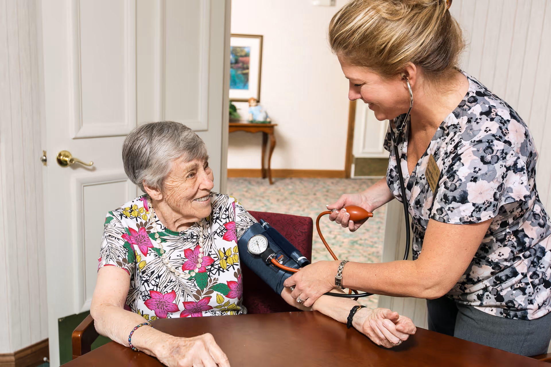 A healthcare worker is smiling and measuring the blood pressure of an elderly woman seated at a table inside a room. The elderly woman is also smiling and wearing a floral shirt. The healthcare worker is using a stethoscope and a blood pressure cuff.