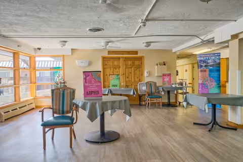 Sunlit common dining/activity room with round tables and chairs, posters on display and large windows.