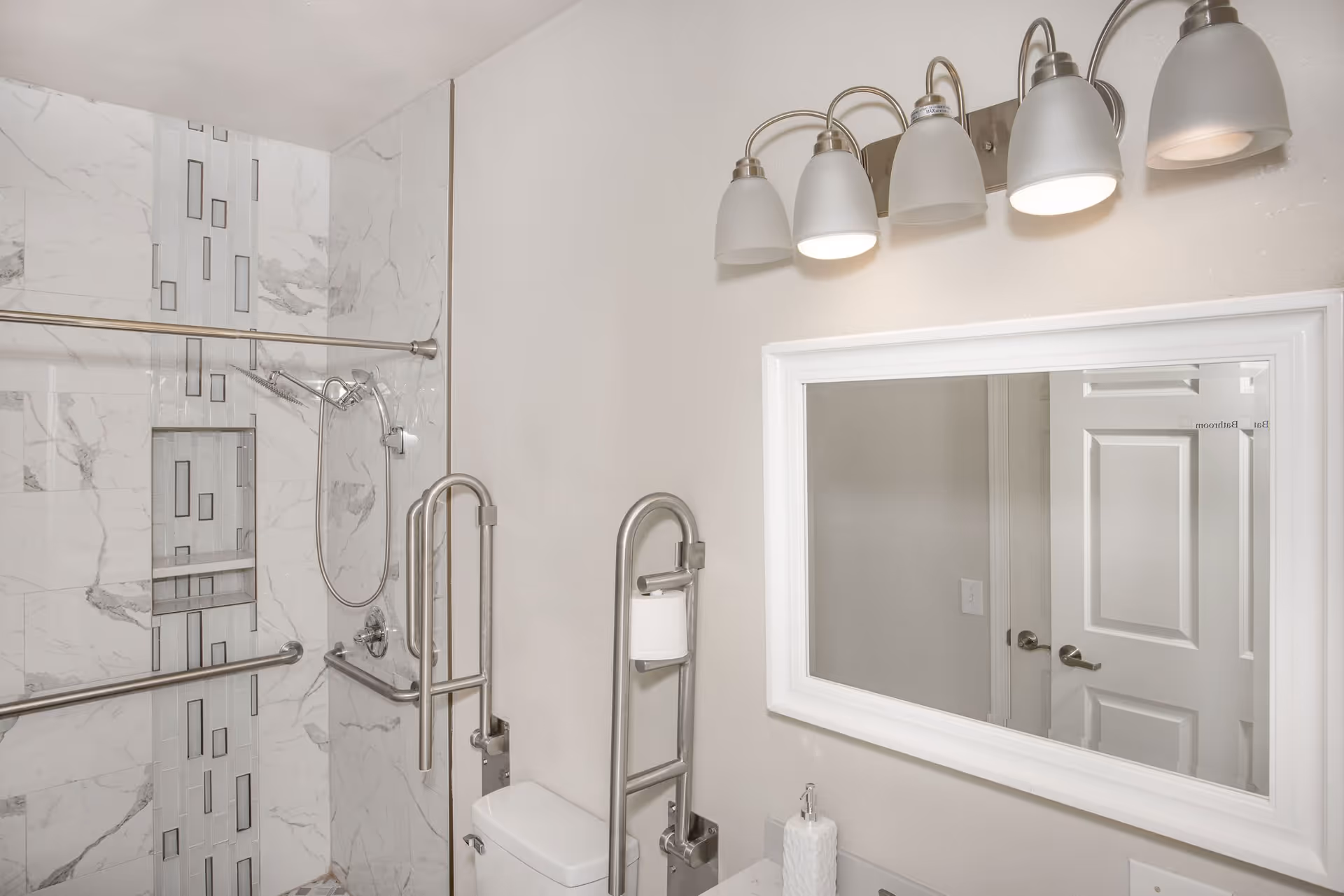 A modern bathroom with a walk-in shower featuring marble-patterned tiles and a vertical decorative strip. The shower has a handheld showerhead and multiple stainless steel grab bars for accessibility. There is a white toilet with a stainless steel grab bar holding a toilet paper roll. Above the sink is a large white-framed mirror reflecting a closed white door. Above the mirror is a light fixture with five frosted glass shades, two of which are lit.