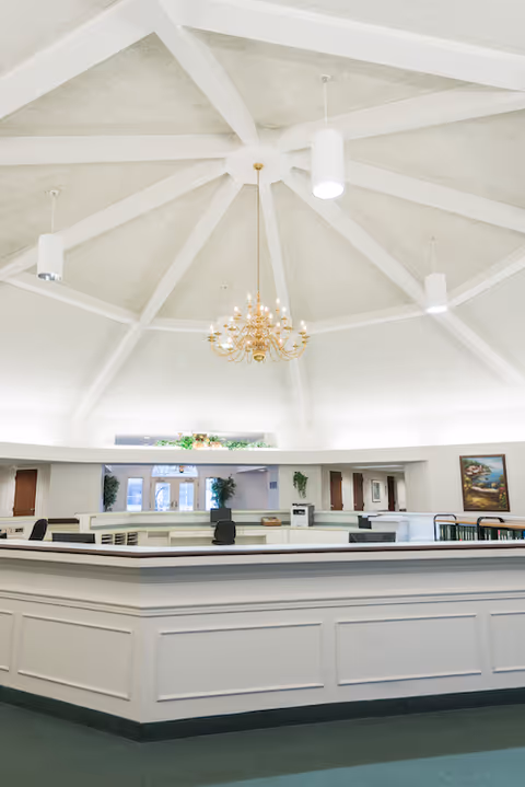 Interior view of a spacious reception or common area with a high vaulted ceiling featuring white beams and a central gold chandelier. The room has a large white reception desk with office chairs behind it, some plants, and framed artwork on the walls.