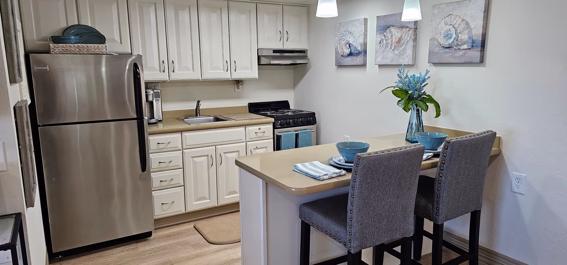 A modern kitchen with white cabinets, a stainless steel refrigerator, a small sink, and a stove with a range hood. A beige countertop extends to form a breakfast bar with two gray upholstered chairs. The bar is set with blue bowls, plates, and napkins. On the wall above the bar are three paintings of seashells, and a vase with blue flowers is placed on the countertop.