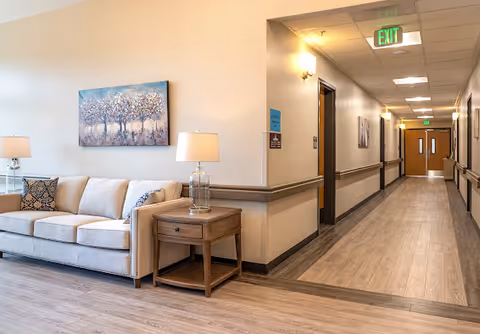 A clean assisted living interior showing a seating area with a sofa, side table and lamp beside a long hallway leading to double doors.