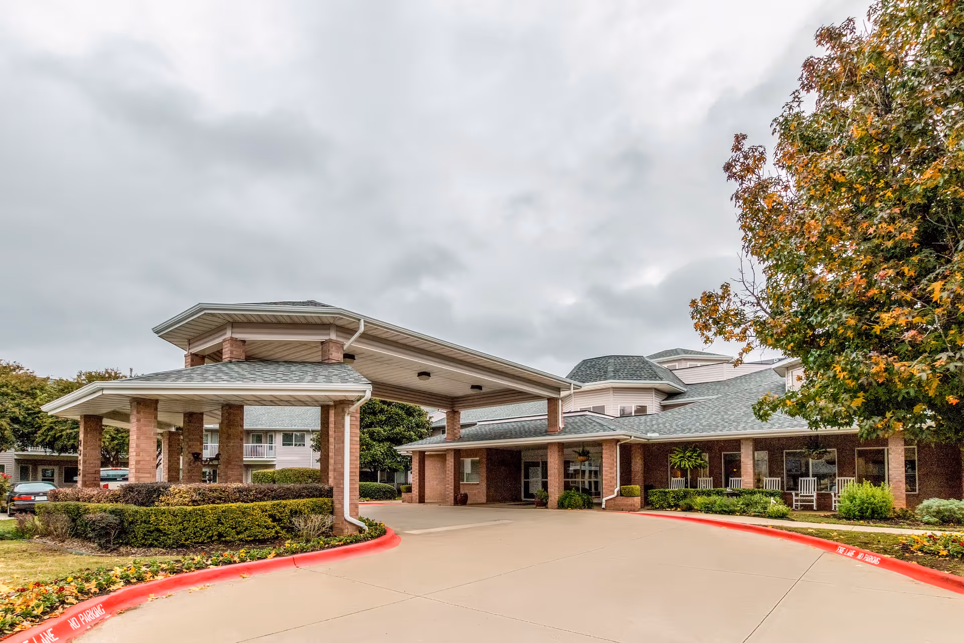 Exterior view of a senior living facility with a covered entrance supported by brick columns, surrounded by landscaped bushes and trees with autumn foliage under a cloudy sky.
