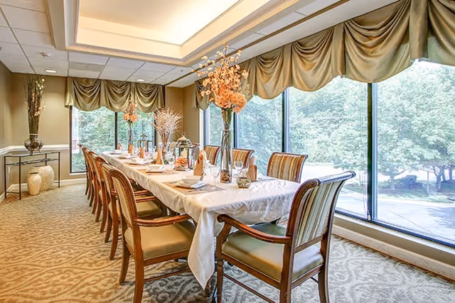 A long dining table set with a white tablecloth, napkins, glassware, and decorative centerpieces in a bright room with large windows and green draped valances, overlooking trees outside.