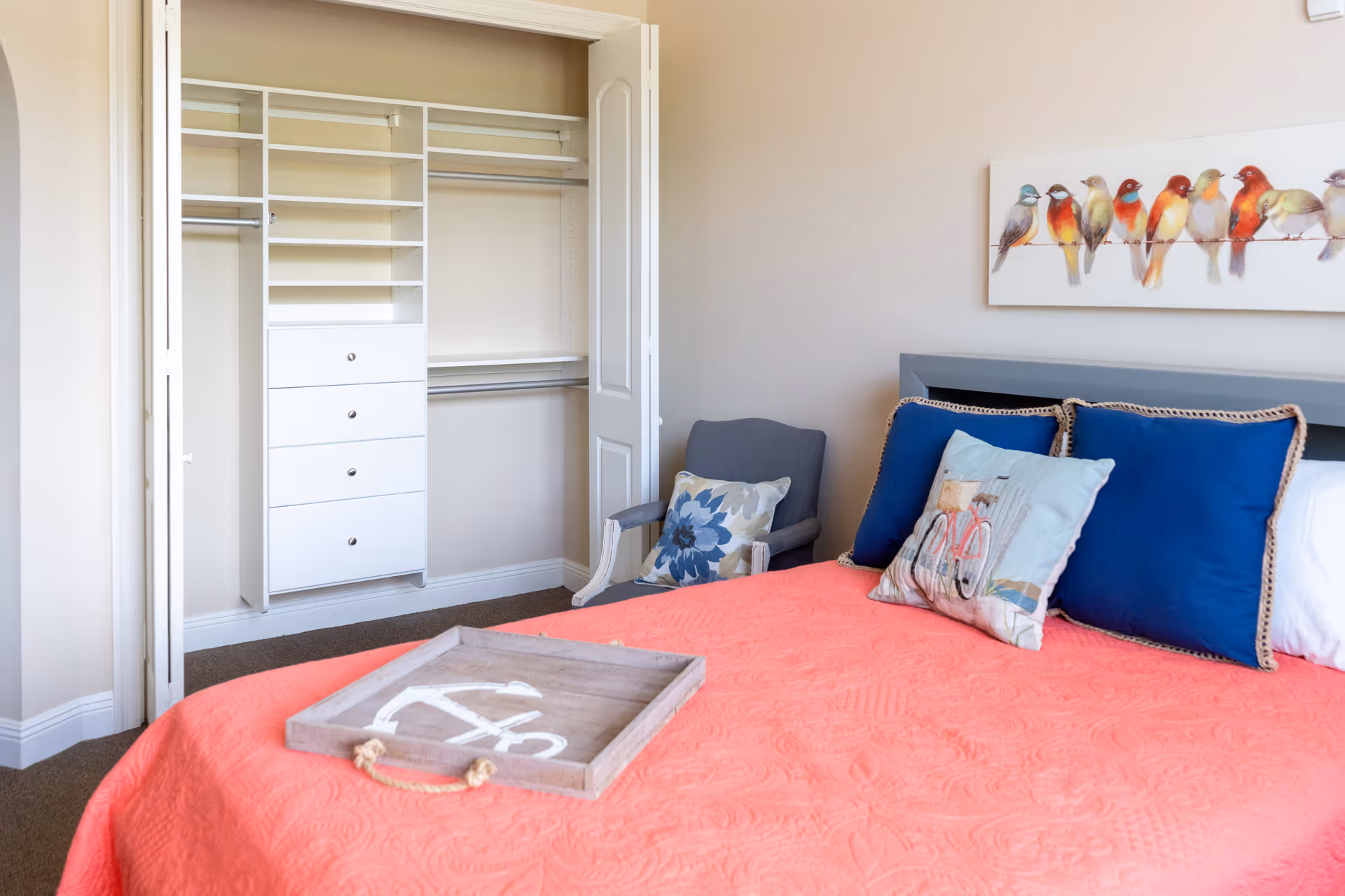A bedroom with a coral-colored bedspread, three decorative pillows, and a wooden tray on the bed. There is a gray armchair with a floral cushion next to an open closet with white shelving and drawers. A painting of colorful birds on a wire hangs on the beige wall above the bed.
