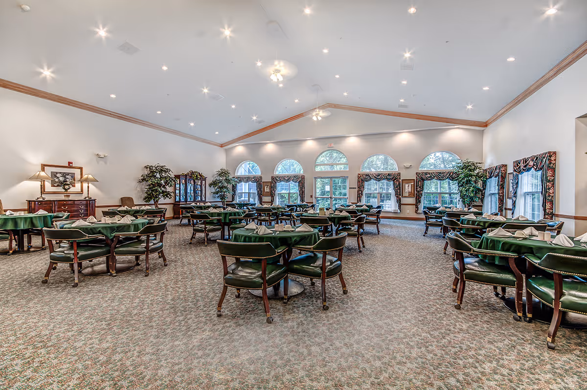 Spacious dining room with multiple round tables covered with green tablecloths and set with folded napkins. The room features large arched windows with patterned curtains, carpeted floor, high ceiling with recessed lighting and ceiling fans, and decorative plants and furniture along the walls.