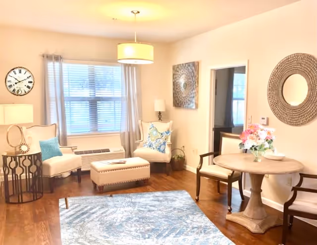 A bright living room with wooden floors, a blue patterned rug, two beige armchairs with decorative pillows, a matching ottoman, a round wooden table with two chairs, a vase of flowers on the table, a large round mirror on the wall, a window with light curtains, and a wall clock.