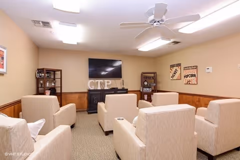 A cozy living room area with six beige armchairs arranged in two rows facing a wall-mounted flat-screen TV. The room has beige walls with wood paneling on the lower half, two small display shelves on either side of a black electric fireplace, and decorative signs on the wall. A ceiling fan and recessed lighting are visible on the ceiling.
