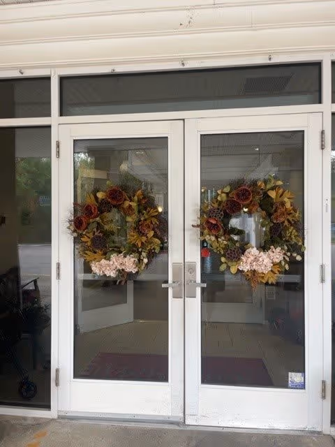 Double glass doors decorated with matching autumn-themed wreaths featuring brown, yellow, and pink flowers. The doors lead into a building interior with visible ceiling lights and a tiled floor.