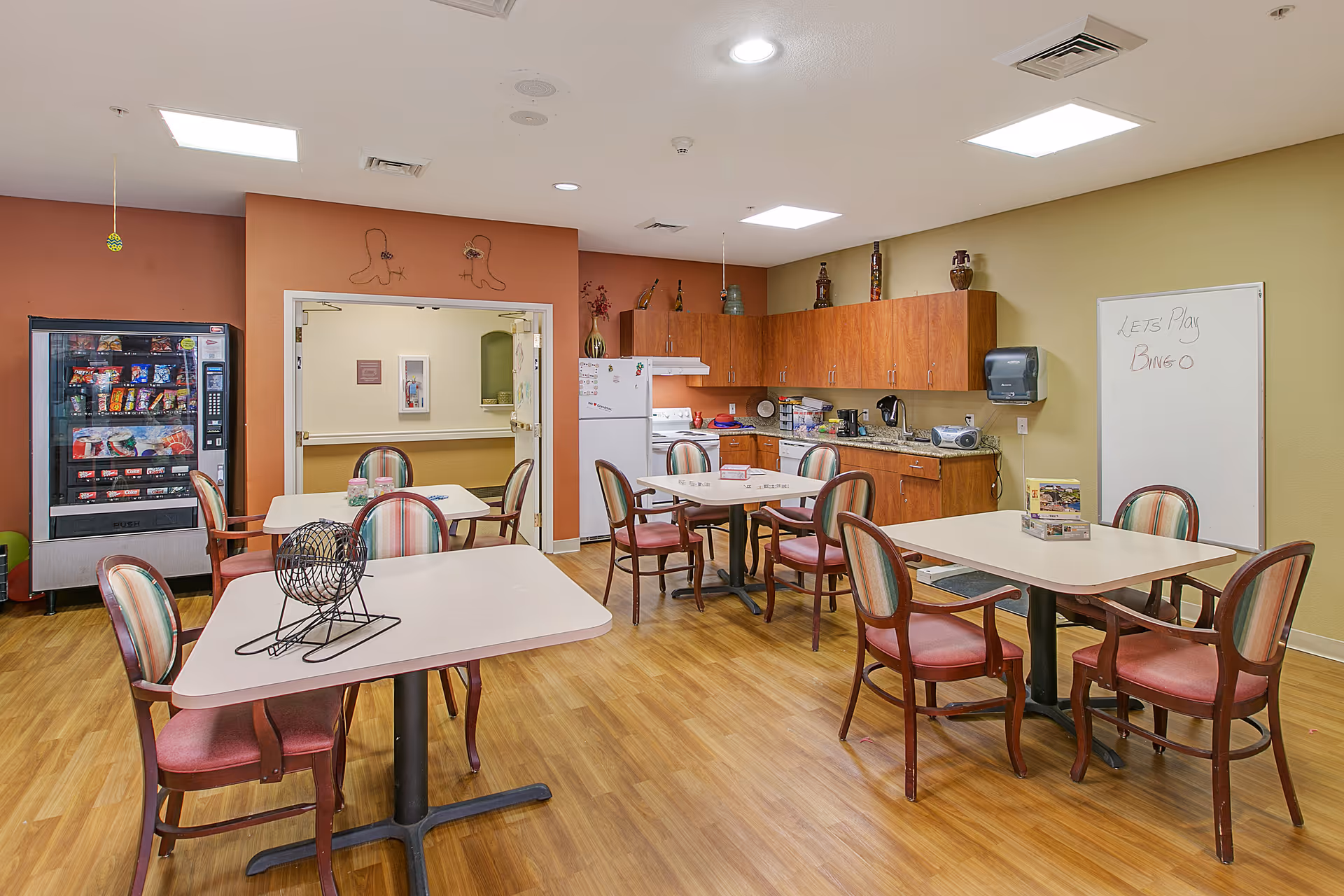 A communal room with several tables and chairs arranged for group activities or dining. The room features a kitchenette with wooden cabinets, a refrigerator, a microwave, and a coffee maker. A vending machine is visible on the left side, and a whiteboard on the right side has the message 'Let's Play Bingo' written on it. The floor is wooden, and the walls are painted in warm tones.