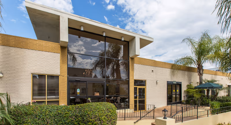 Exterior view of Alexandria Care Center showing a single-story building with large glass windows, beige brick walls, palm trees, and a clear blue sky with some clouds.