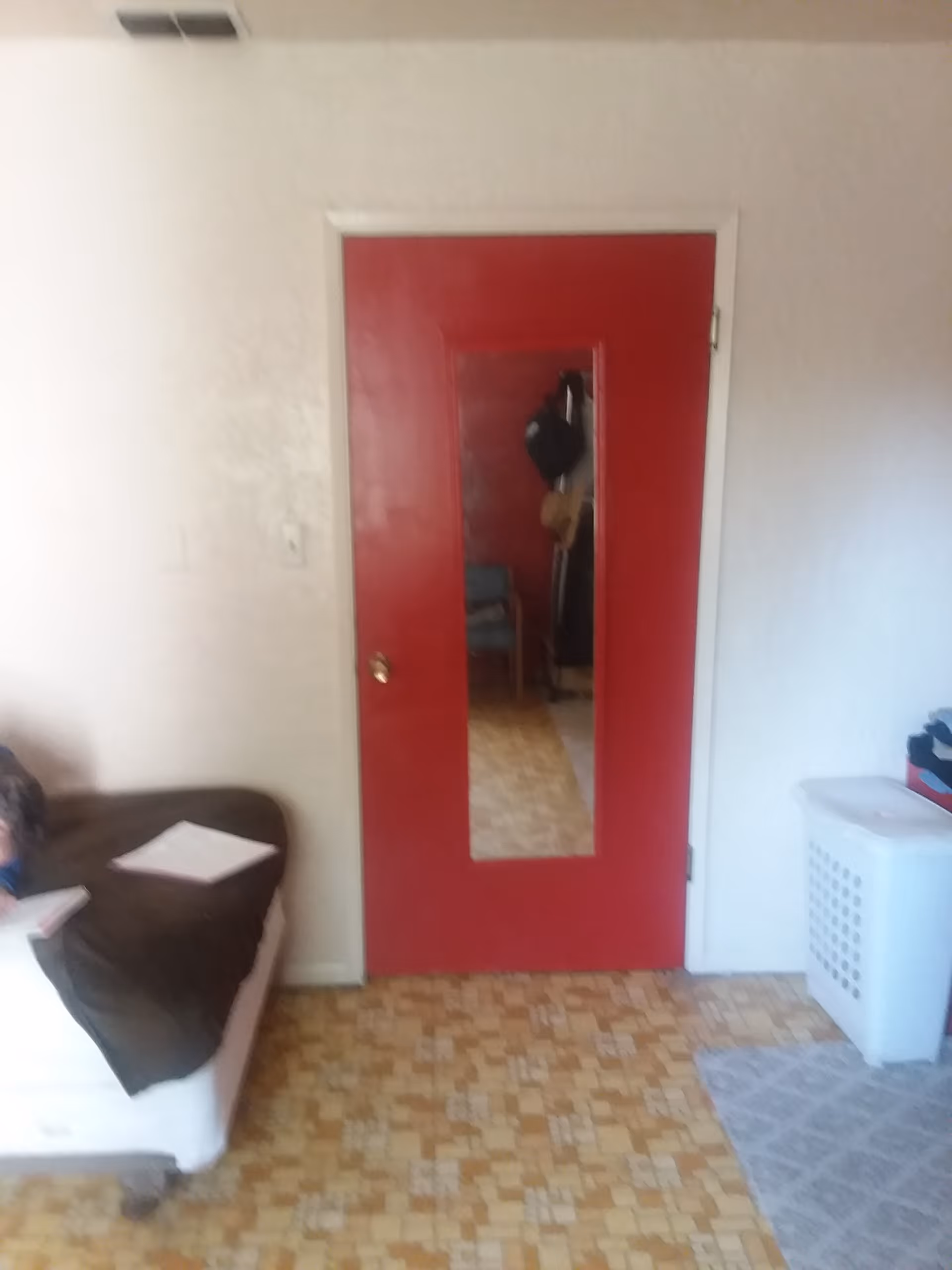 Interior view of a room with a red door featuring a rectangular mirror in the center. To the left, there is a white couch with a dark brown blanket and a piece of paper on it. To the right, there is a white laundry basket and a gray patterned rug on a tiled floor.