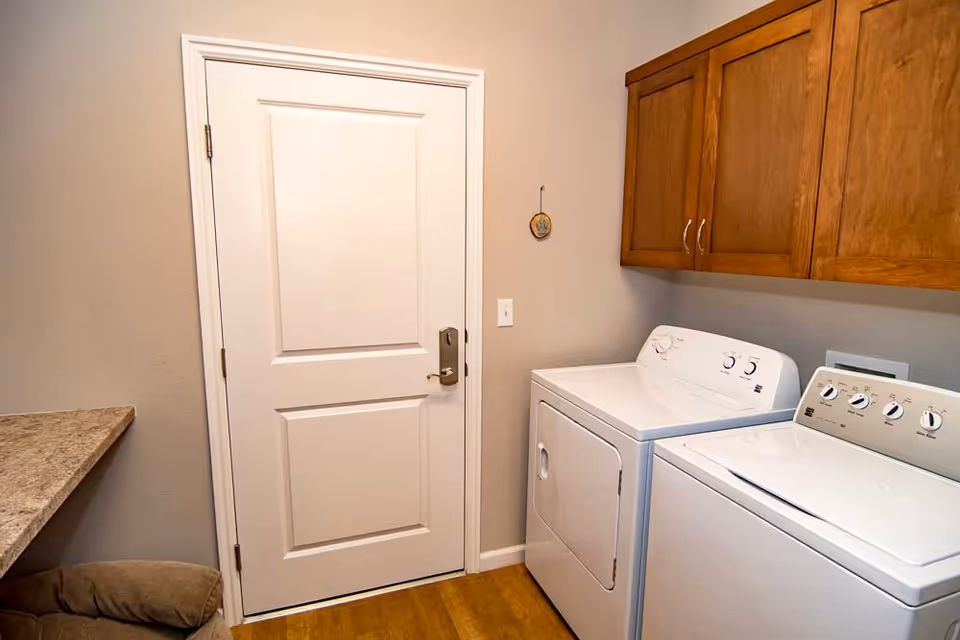 Laundry room with a white washing machine and dryer side by side, wooden cabinets above them, a white door with a silver handle, and a countertop on the left side.
