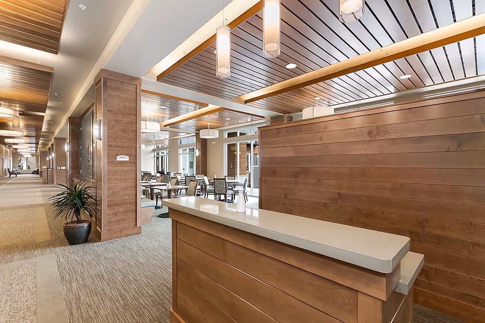 Interior view of a senior living facility featuring a reception desk in the foreground with wood paneling and a light-colored countertop. Behind the desk is a dining area with multiple tables and chairs, illuminated by modern pendant lights and natural light from large windows. The space has a warm, inviting atmosphere with wood accents on the ceiling and walls, and a long hallway extending to the left with additional seating and lighting fixtures.