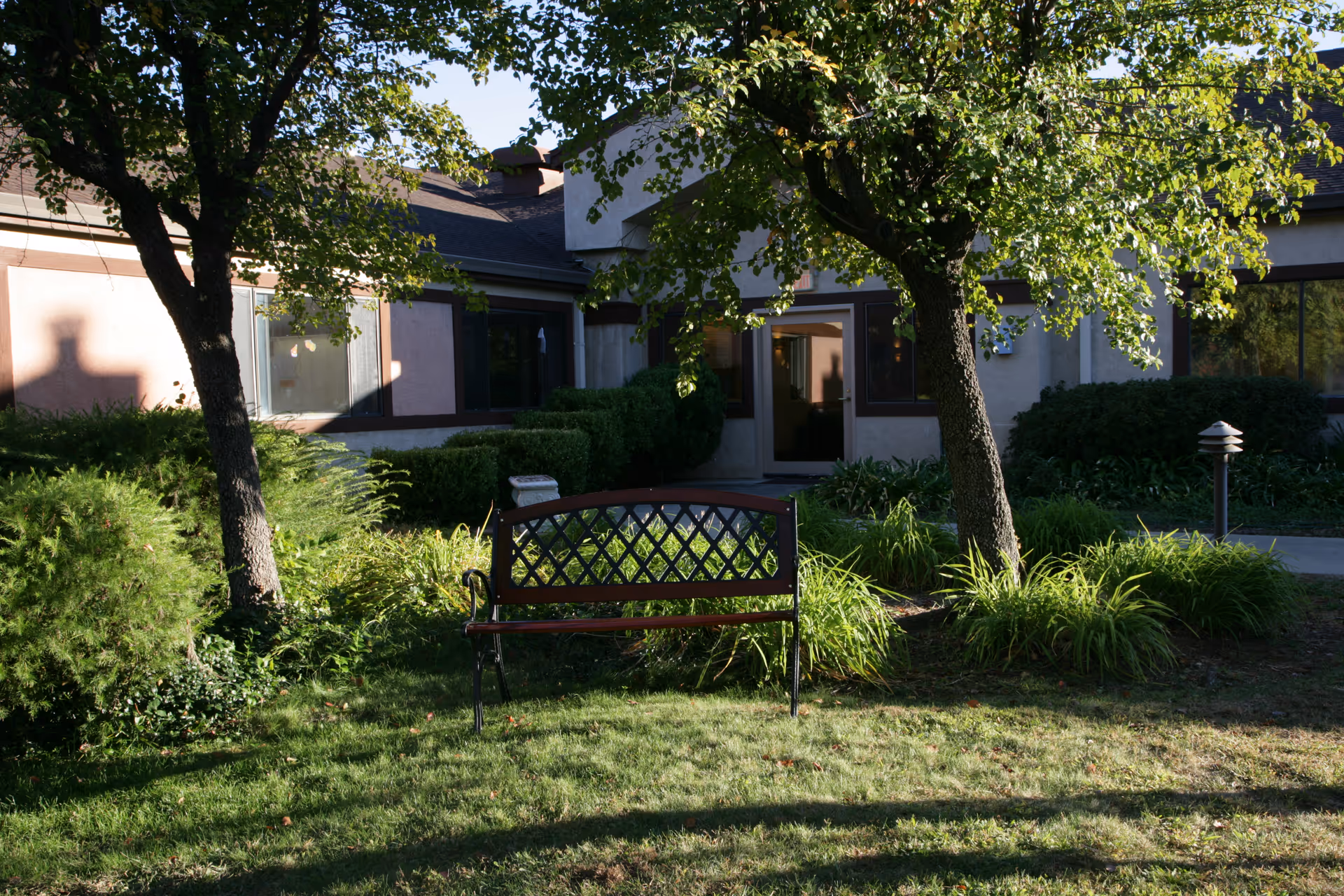 A bench sits in a landscaped courtyard with trees in front of a one-story senior living building.