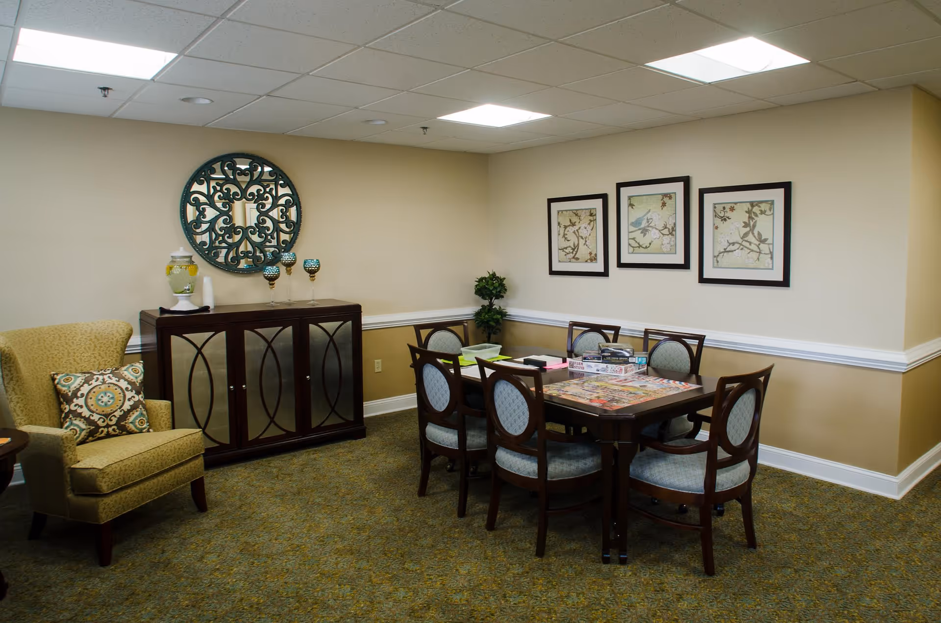 A cozy interior room with a wooden table surrounded by six upholstered chairs. On the table, there are board games and puzzles. To the left, there is a cushioned armchair with a decorative pillow. Against the wall, there is a dark wooden cabinet with a decorative round mirror above it and three decorative goblets. The walls are painted beige with white trim, and three framed pictures featuring birds and floral designs hang on the wall. The room is carpeted and lit by ceiling lights.
