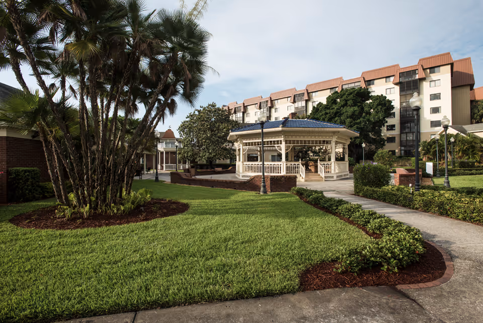 Outdoor garden area with a paved walkway, green grass, palm trees, and a white gazebo with a blue roof. In the background, there is a multi-story building with a reddish-brown roof and several windows. The sky is partly cloudy.