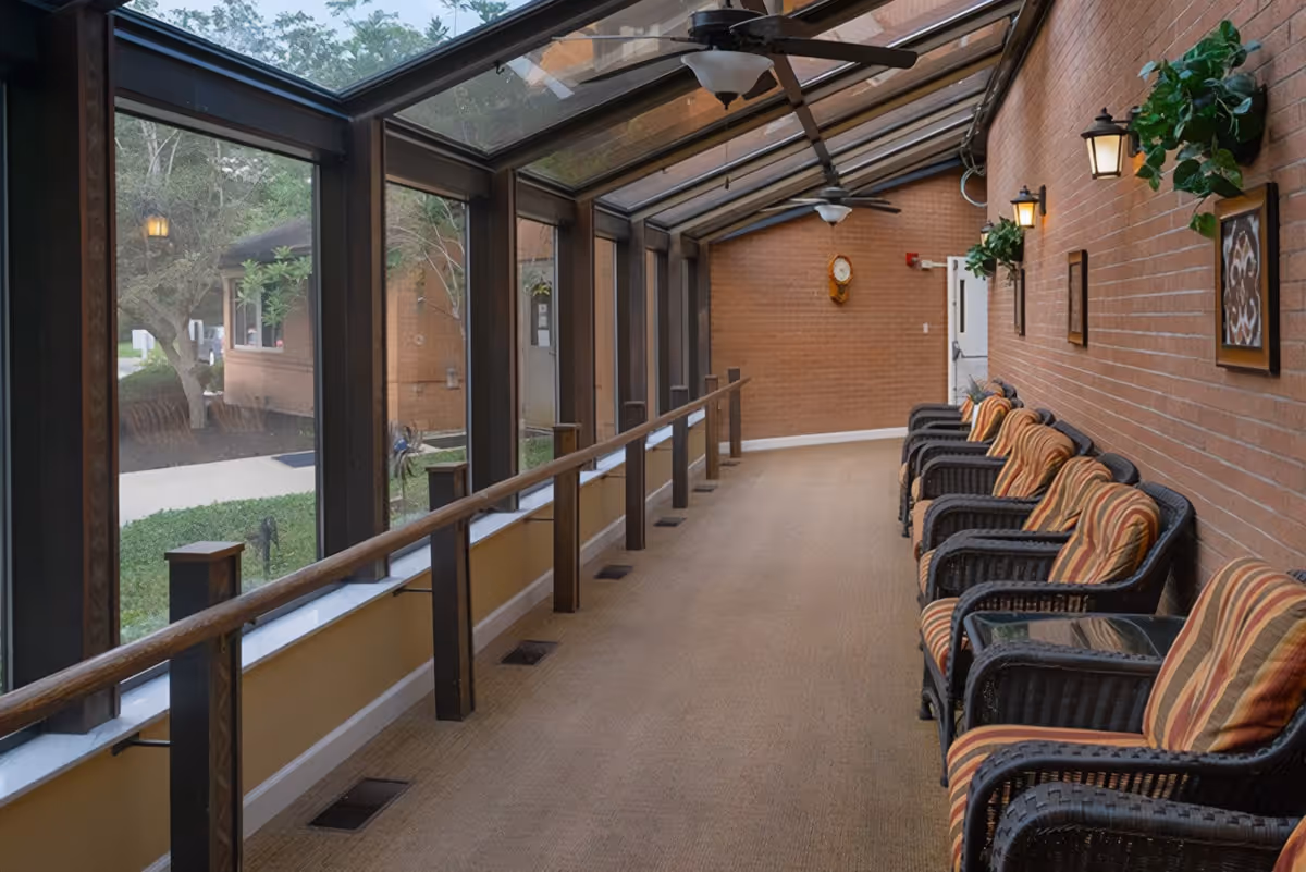 A long indoor hallway with large windows and a glass ceiling on one side, overlooking an outdoor garden area. The opposite wall is brick with several wall-mounted lights, framed artwork, and green plants. There are multiple cushioned wicker chairs lined up along the brick wall, and ceiling fans are mounted on the ceiling.