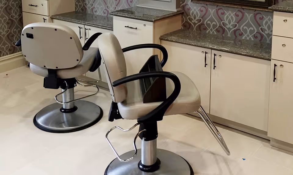 Interior view of a salon area with two beige salon chairs in front of a counter with granite countertops and cabinets. The wall behind the counter has a decorative patterned wallpaper.