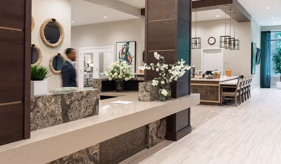 Bright modern senior living reception and dining area with a marble front desk, floral arrangements, seating, and a blurred staff member.