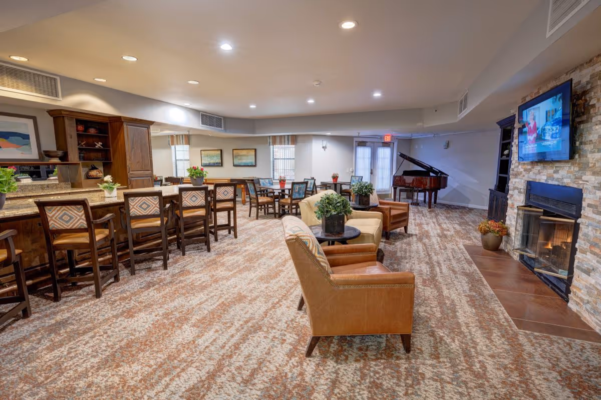 A spacious senior living facility common area with a carpeted floor, several armchairs and small tables with potted plants, a stone fireplace with a TV mounted above it, a grand piano in the corner, and a dining area with tables and chairs near large windows.